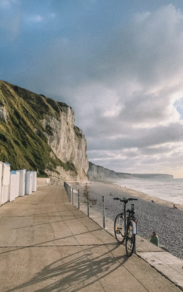Bike on a walkway by tall cliffs and a pebbled beach with ocean views under a cloudy sky.